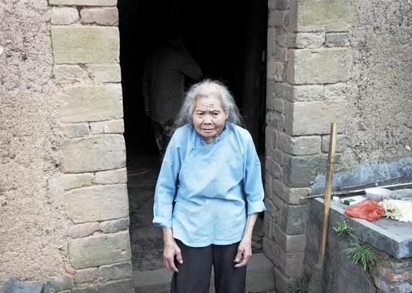 蒋爱招老人站在家门口,家里居住的泥土房子,生活困难。