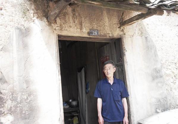 叶天金老人站在家门口,家里居住泥土房子,墙壁多处裂缝,生活条件艰苦。